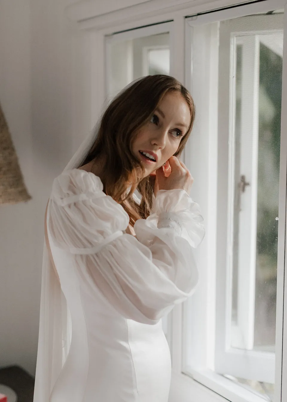 Elyse fastening her earring in soft window light. At Fig Tree Villa in Ewingsdale, Byron Bay, for an Elopement Collective elopement. Photograph by Bulb Creative.