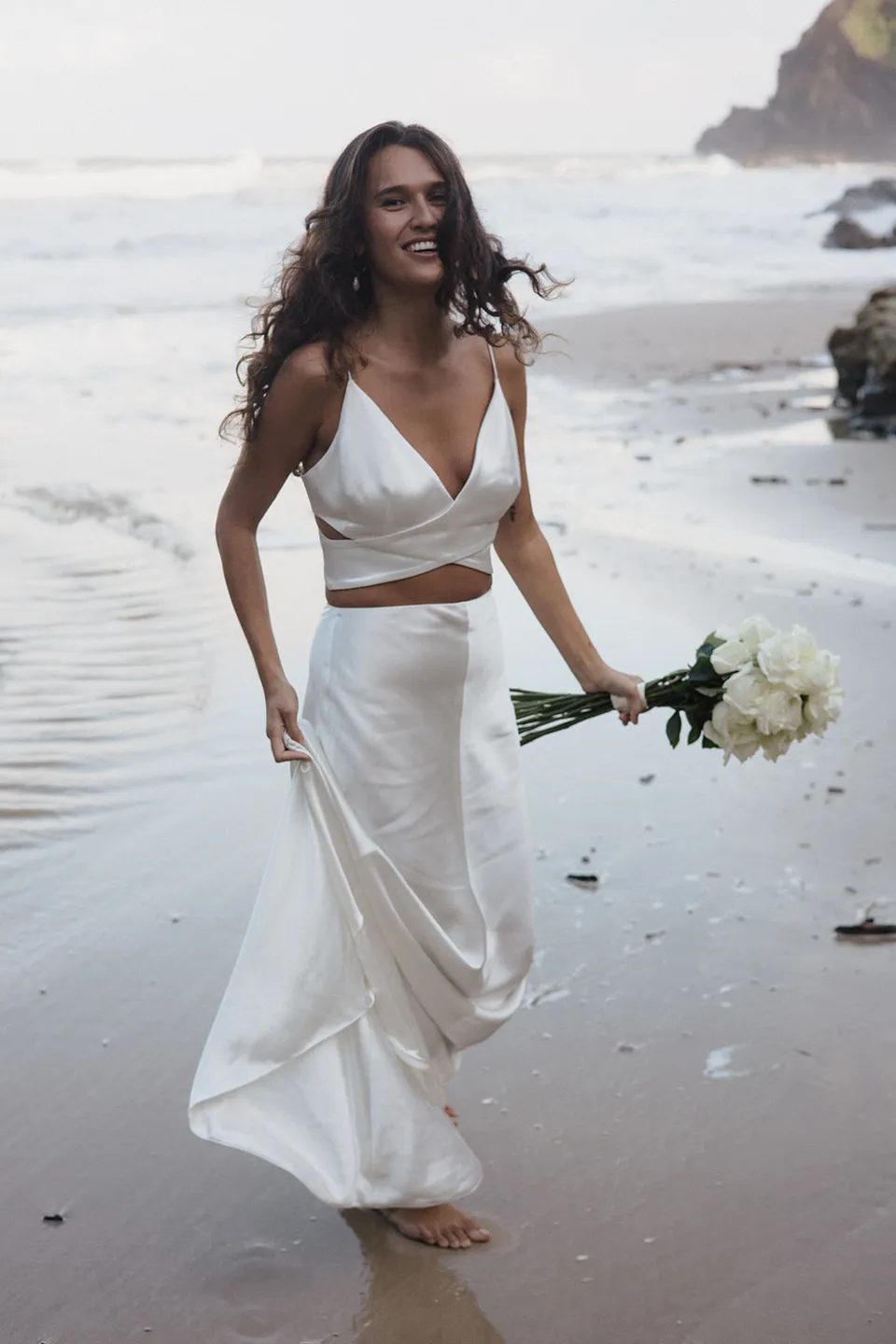 Rachel walking barefoot along the shoreline with her white bouquet, with flowers by Fifth Avenue Florist. At Whites Beach, Broken Head, near Byron Bay, for an Elopement Collective elopement. Photograph by Jason Lucas.