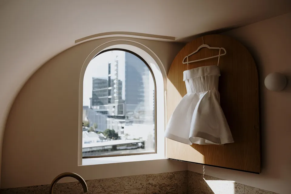 Danielle’s short white dress hanging beside an arched window overlooking Brisbane, at The Calile Hotel in Fortitude Valley, Brisbane, for an Elopement Collective elopement. Photo by George Bowden.