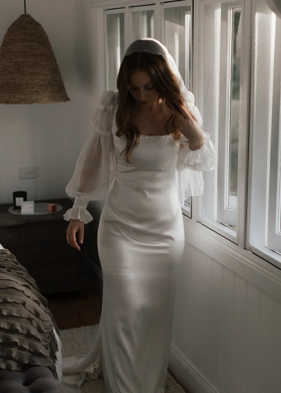 Elyse looking down at her dress beside the window. At Fig Tree Villa in Ewingsdale, Byron Bay, for an Elopement Collective elopement. Photograph by Bulb Creative.