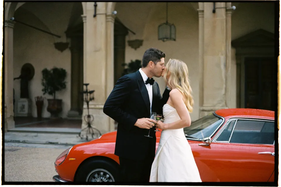 Holly and Matthew holding hands and kissing beside the red vintage sports car, at Villa San Michele in Fiesole above Florence, Tuscany, Italy, for an Elopement Collective elopement. Film photograph by Khara from House of Lucie.