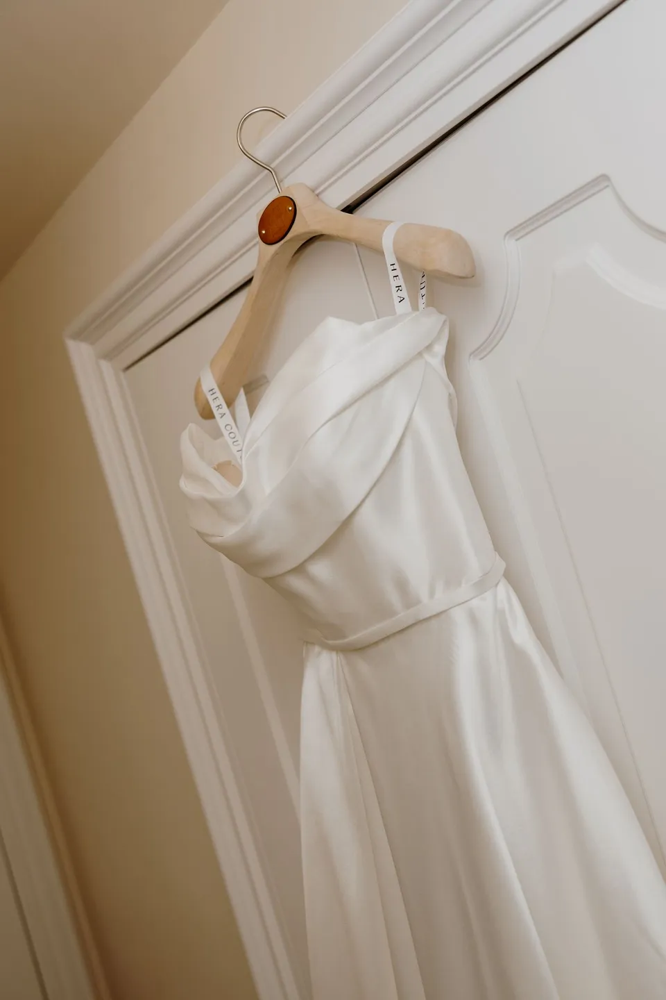 Zoe’s white wedding dress with a draped neckline hanging from a wooden hanger against a white door, at Passalacqua in Moltrasio, Lake Como, Italy, for an Elopement Collective elopement. Photo by George Bowden.
