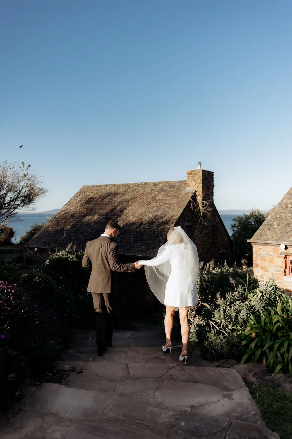 Kaitlyn and Russell holding hands as they walk down a stone path between the cottages. At Piermont Retreat on Tasmania’s east coast, for an Elopement Collective elopement. Photograph by Omar of By The Wilde.