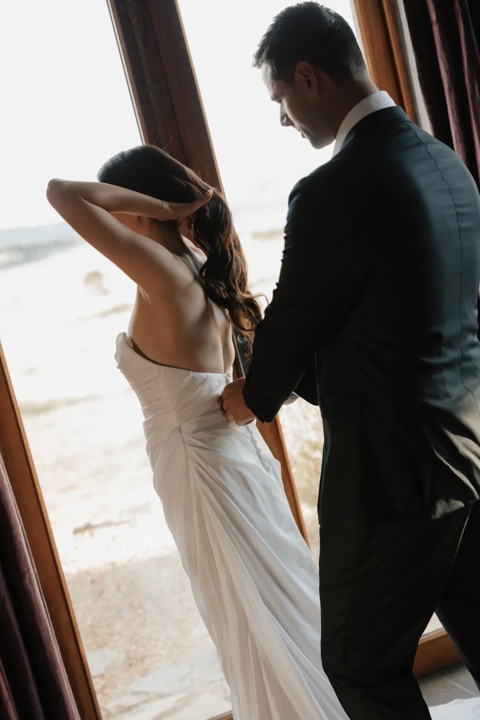 Anthony helping Dani with her white wedding dress beside a bright window, at Thalia Haven on Tasmania’s east coast, for an Elopement Collective elopement. Photograph by Bulb Creative.