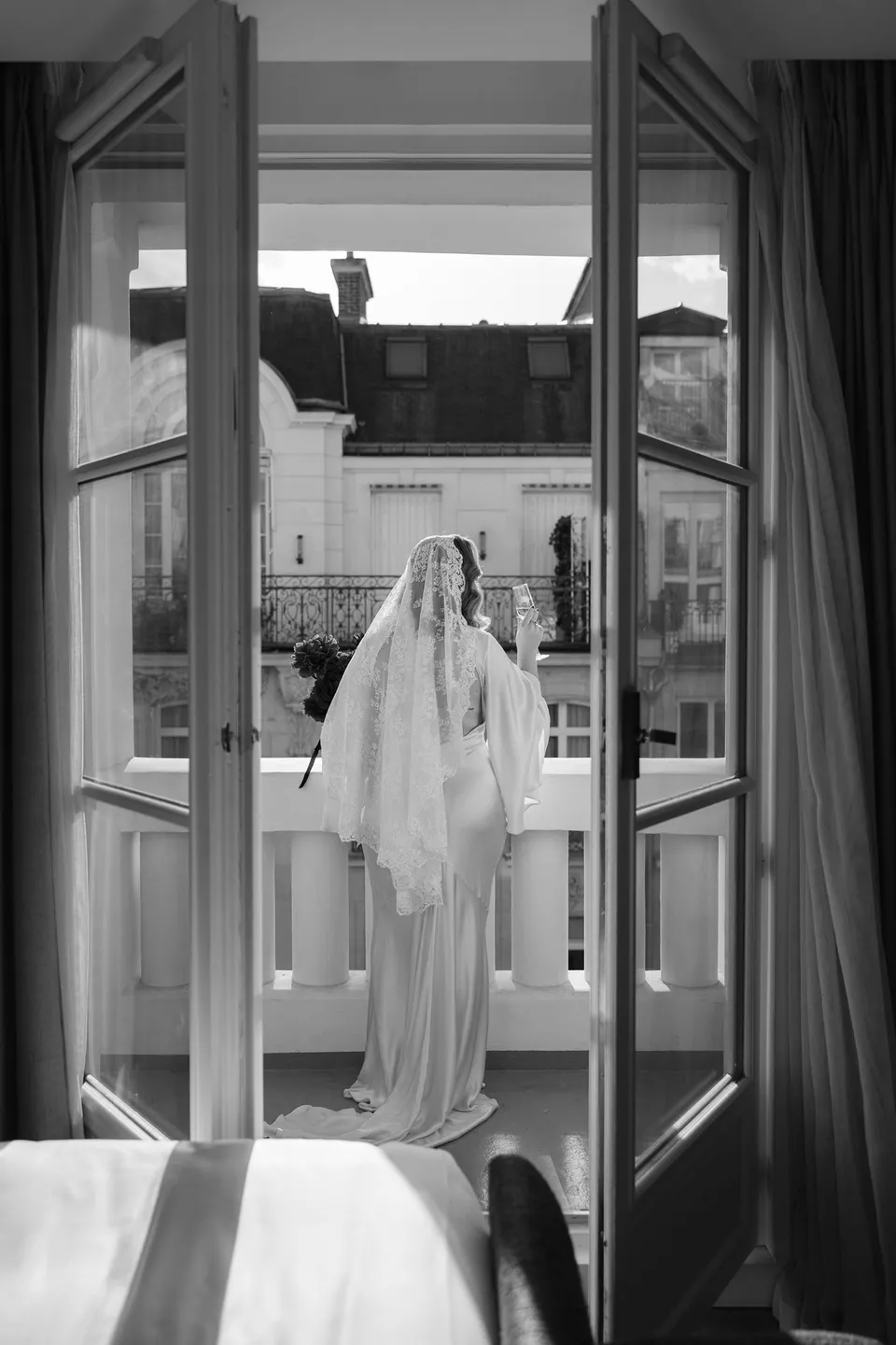 Black and white view of Kara standing on the balcony through open French doors, with the hotel bed in the foreground, at Hotel Marignan Champs-Élysées in Paris, France, during an Elopement Collective elopement. Photograph by George Bowden.