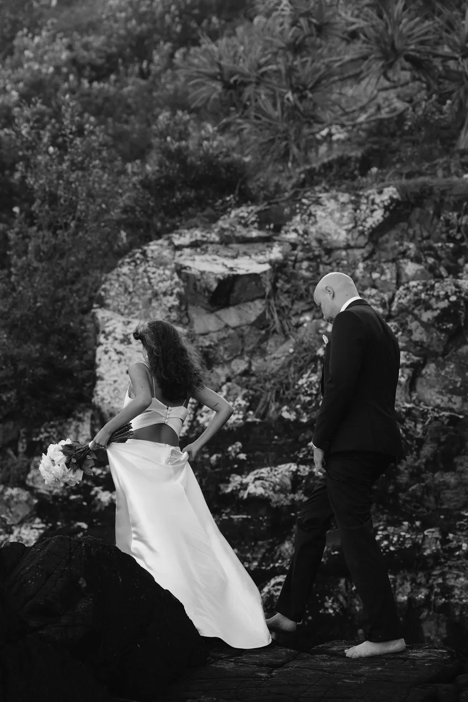 Black-and-white photograph of Rachel and Dane climbing the rocks with their backs to the camera. At Whites Beach, Broken Head, near Byron Bay, for an Elopement Collective elopement. Photograph by Jason Lucas.