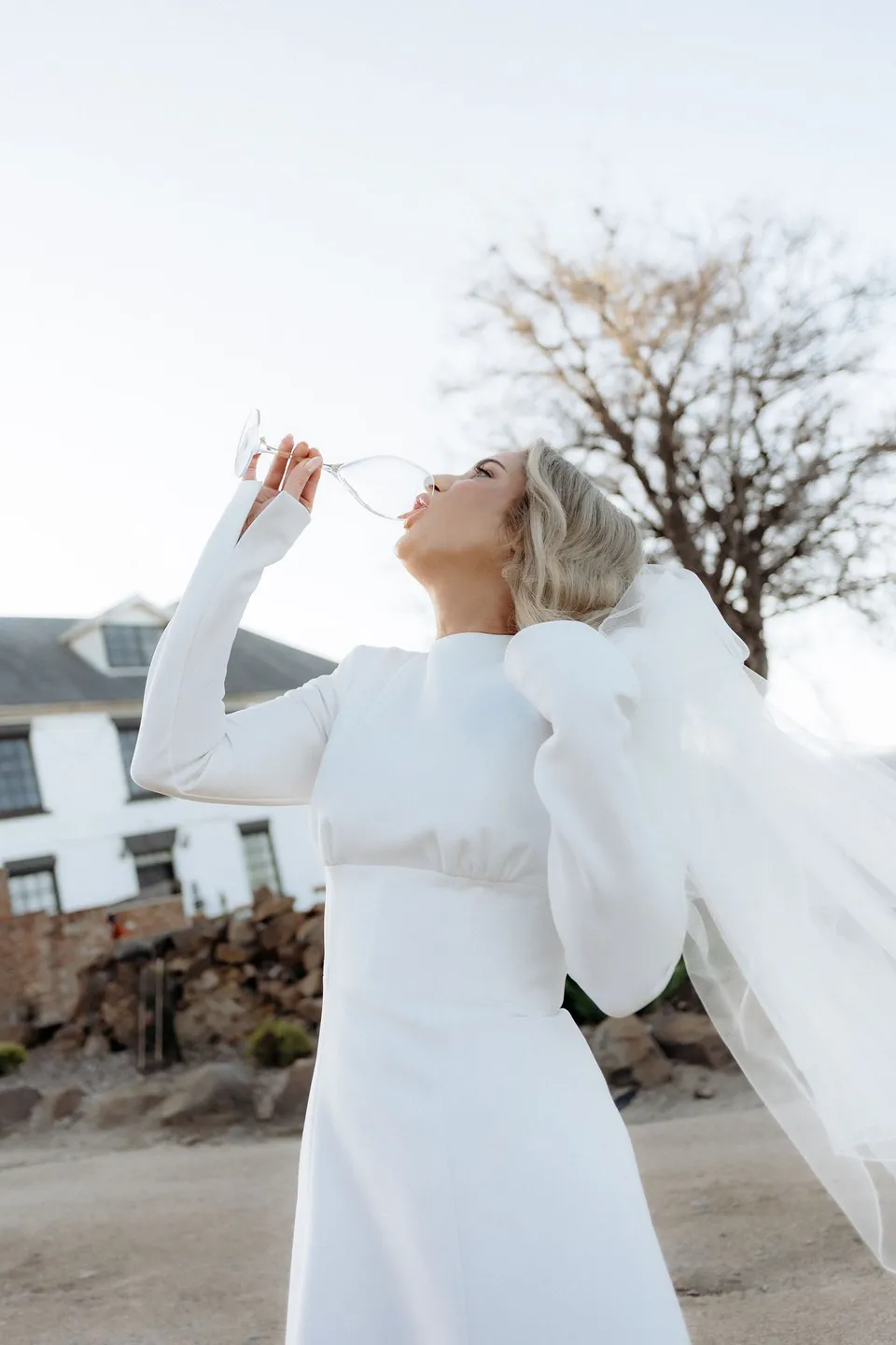 Kaitlyn sipping from a stemmed glass outside the white homestead, with her veil catching the light. At Piermont Retreat on Tasmania’s east coast, for an Elopement Collective elopement. Photograph by Omar of By The Wilde.