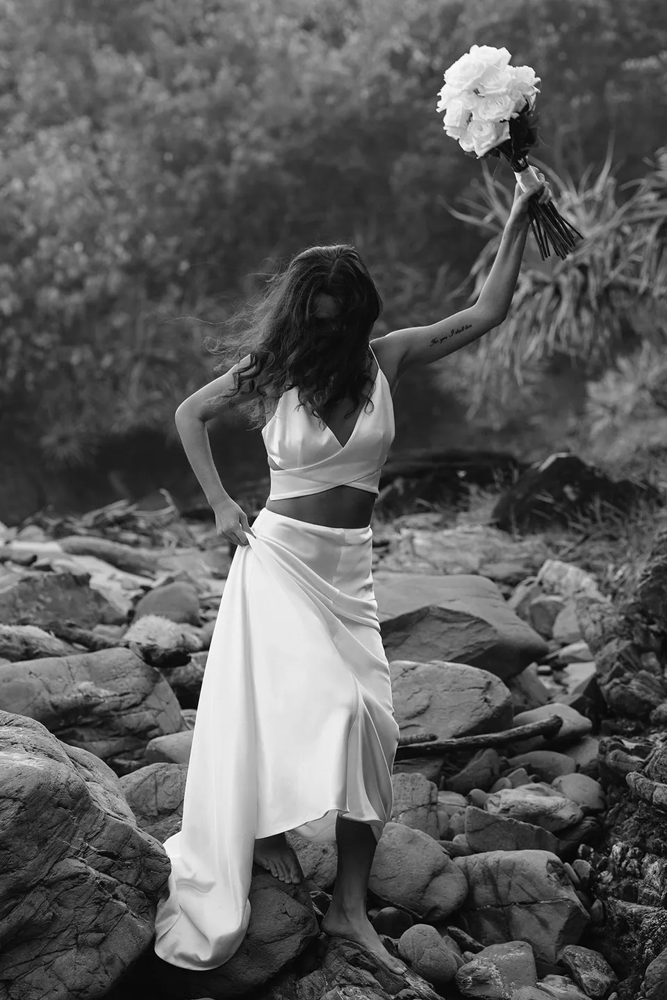 Black-and-white portrait of Rachel raising her bouquet beside the rock face, with flowers by Fifth Avenue Florist. At Whites Beach, Broken Head, near Byron Bay, for an Elopement Collective elopement. Photograph by Jason Lucas.