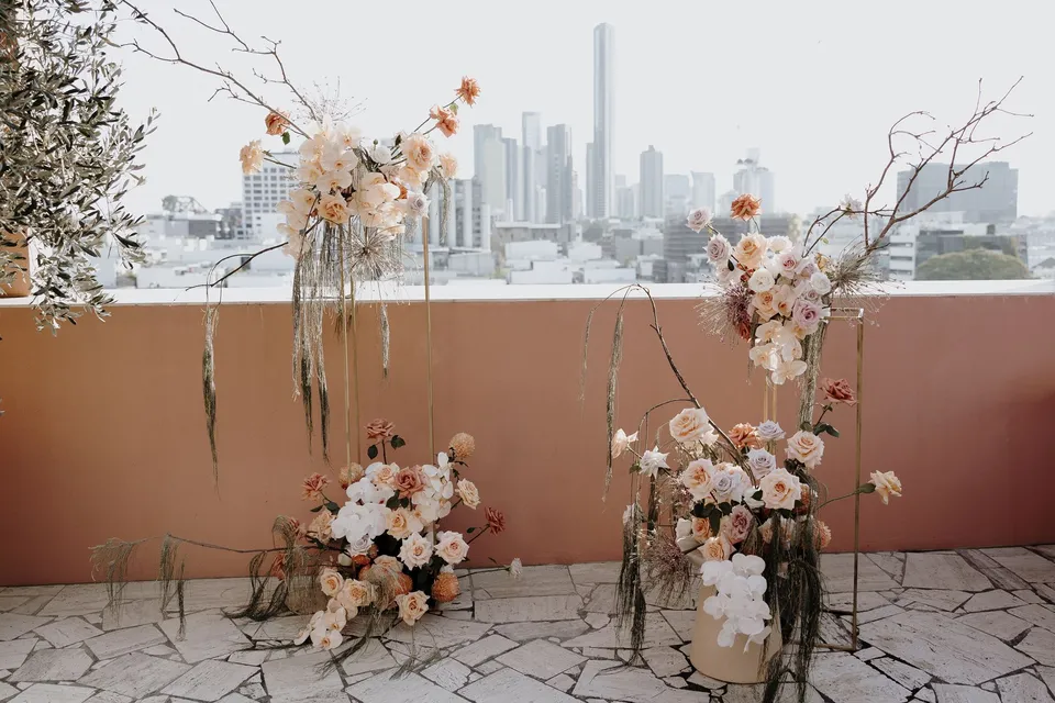 Peach and white ceremony flowers by Mrs Gibbons Florals arranged among bare branches on a pink terrace overlooking Brisbane, at The Calile Hotel in Fortitude Valley, Brisbane, for an Elopement Collective elopement. Photo by George Bowden.