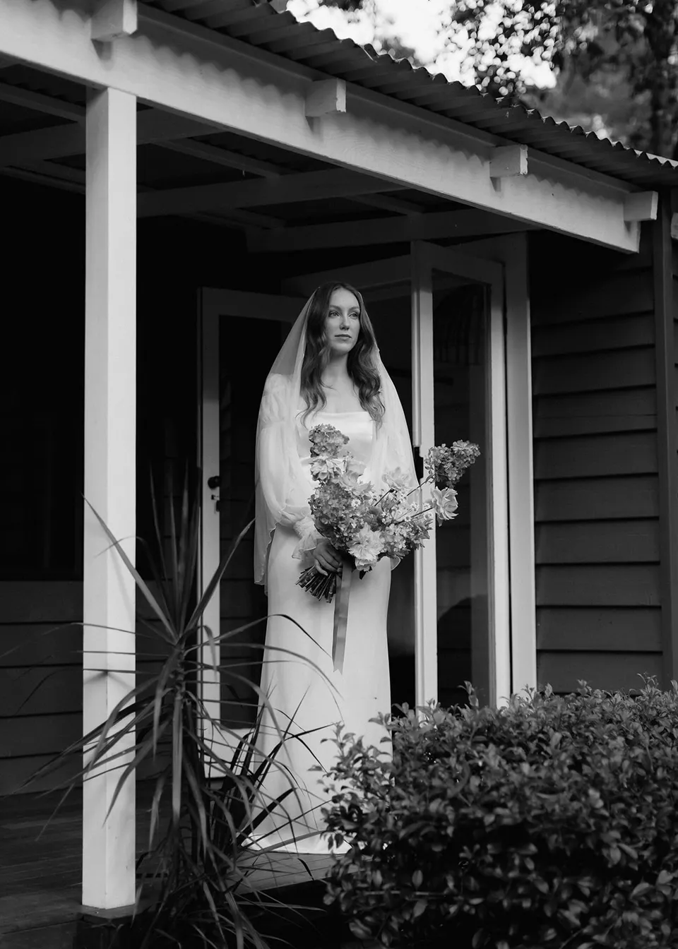 Black-and-white photograph of Elyse on the verandah beneath the roofline. At Fig Tree Villa in Ewingsdale, Byron Bay, for an Elopement Collective elopement. Photograph by Bulb Creative.