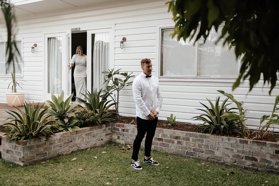 Laura stepping out of the white beach house as Matt waits in the garden, at Grounds of Cabarita in Cabarita Beach, NSW, for an Elopement Collective elopement. Photo by Bulb Creative.