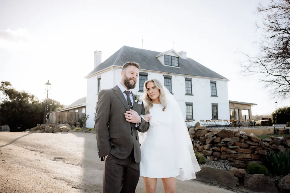 Kaitlyn and Russell standing arm in arm with drinks outside Piermont’s white homestead. At Piermont Retreat on Tasmania’s east coast, for an Elopement Collective elopement. Photograph by Omar of By The Wilde.