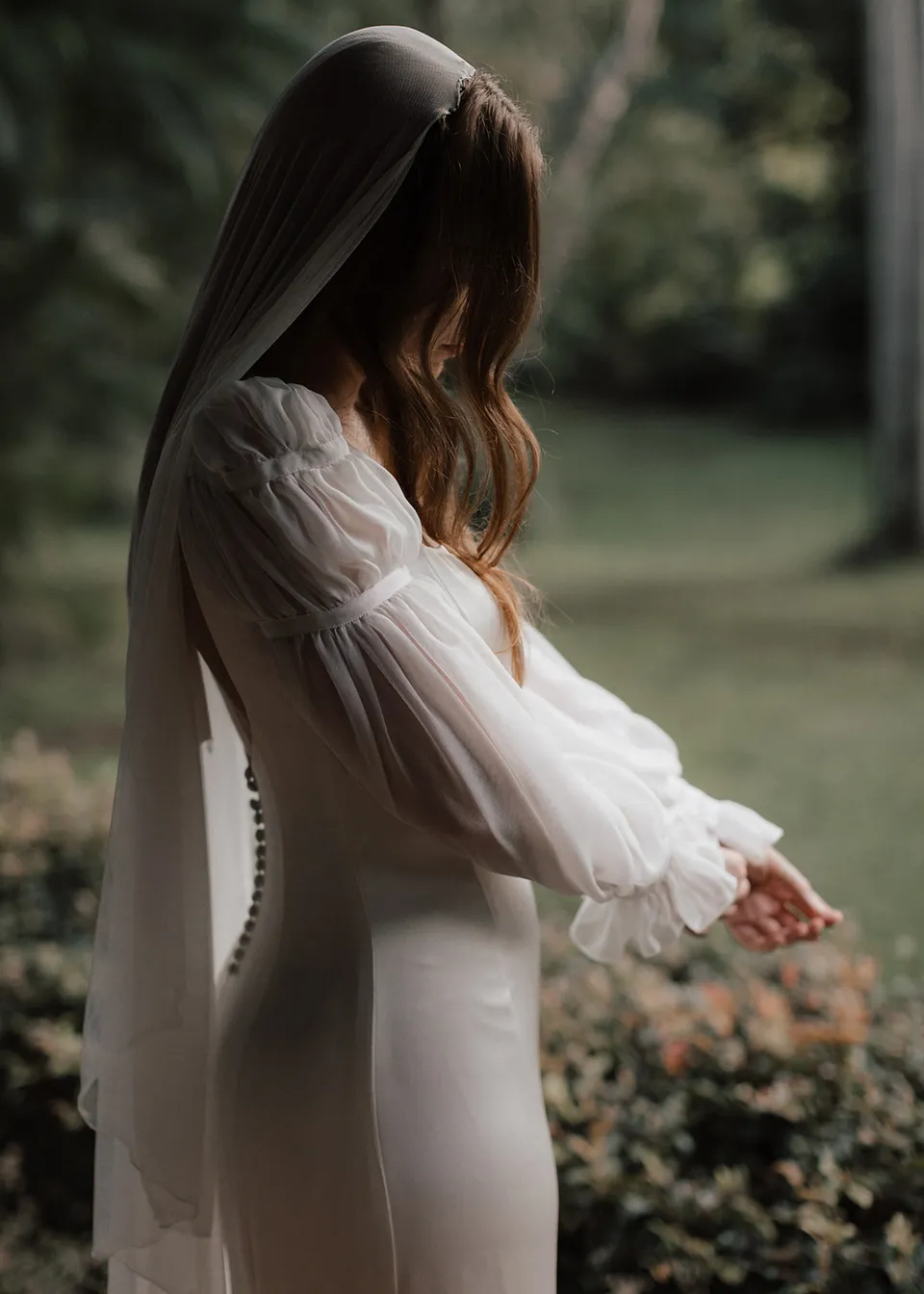 Elyse looking down at her sleeves beside the garden. At Fig Tree Villa in Ewingsdale, Byron Bay, for an Elopement Collective elopement. Photograph by Bulb Creative.