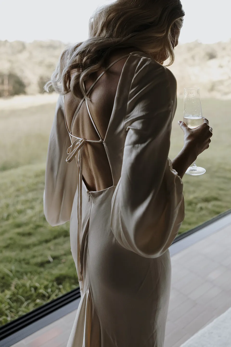 Gabrielle holding champagne beside the cabin window, showing the open back and tie detail of her Shona Joy dress, at Roundhill Retreat in Obi Obi, Sunshine Coast Hinterland, Queensland, for an Elopement Collective elopement. Photo by George Bowden.