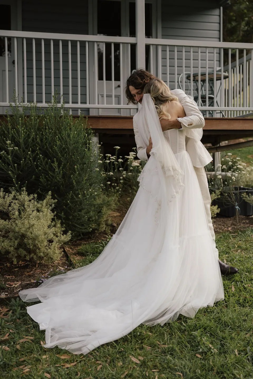 Bridget and James embracing at the foot of the verandah steps, her train spread across the grass. At Cudgera Creek in northern New South Wales, for an Elopement Collective elopement. Photograph by Bulb Creative.