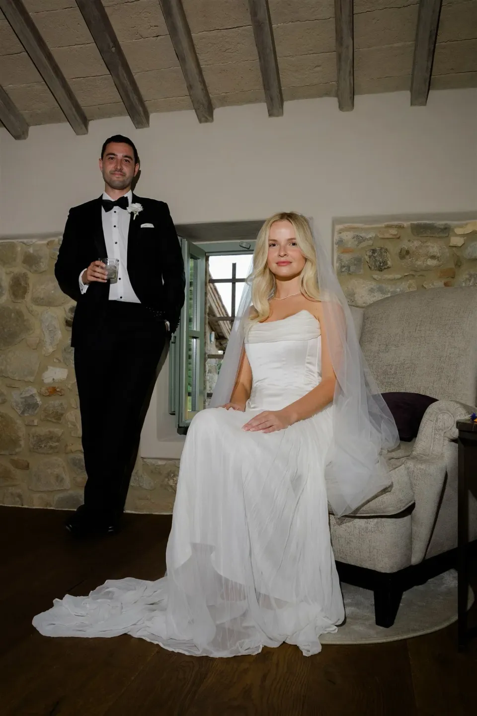 Shelby seated in her white Kate Willa dress while Jack stands beside her holding a drink, in Umbria, Italy, for an Elopement Collective elopement. Photo by James from House of Love.