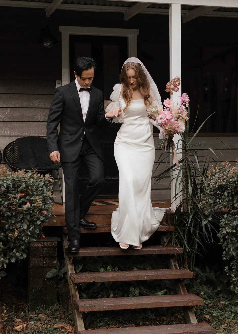 Jonathan helping Elyse down the timber steps while she carries her bouquet, with flowers by The Perfect Party Co. At Fig Tree Villa in Ewingsdale, Byron Bay, for an Elopement Collective elopement. Photograph by Bulb Creative.