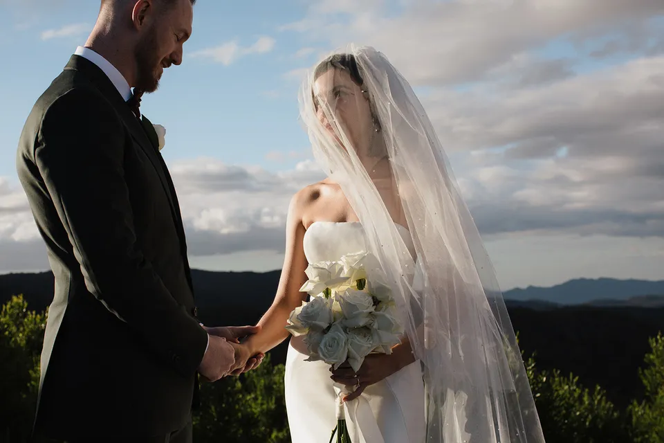 Meg and Ryan holding hands during their ceremony, with her veil and white rose bouquet by The Romantics. At The Keep on Tasmania’s east coast, for an Elopement Collective elopement. Photograph by Jenna Agius.