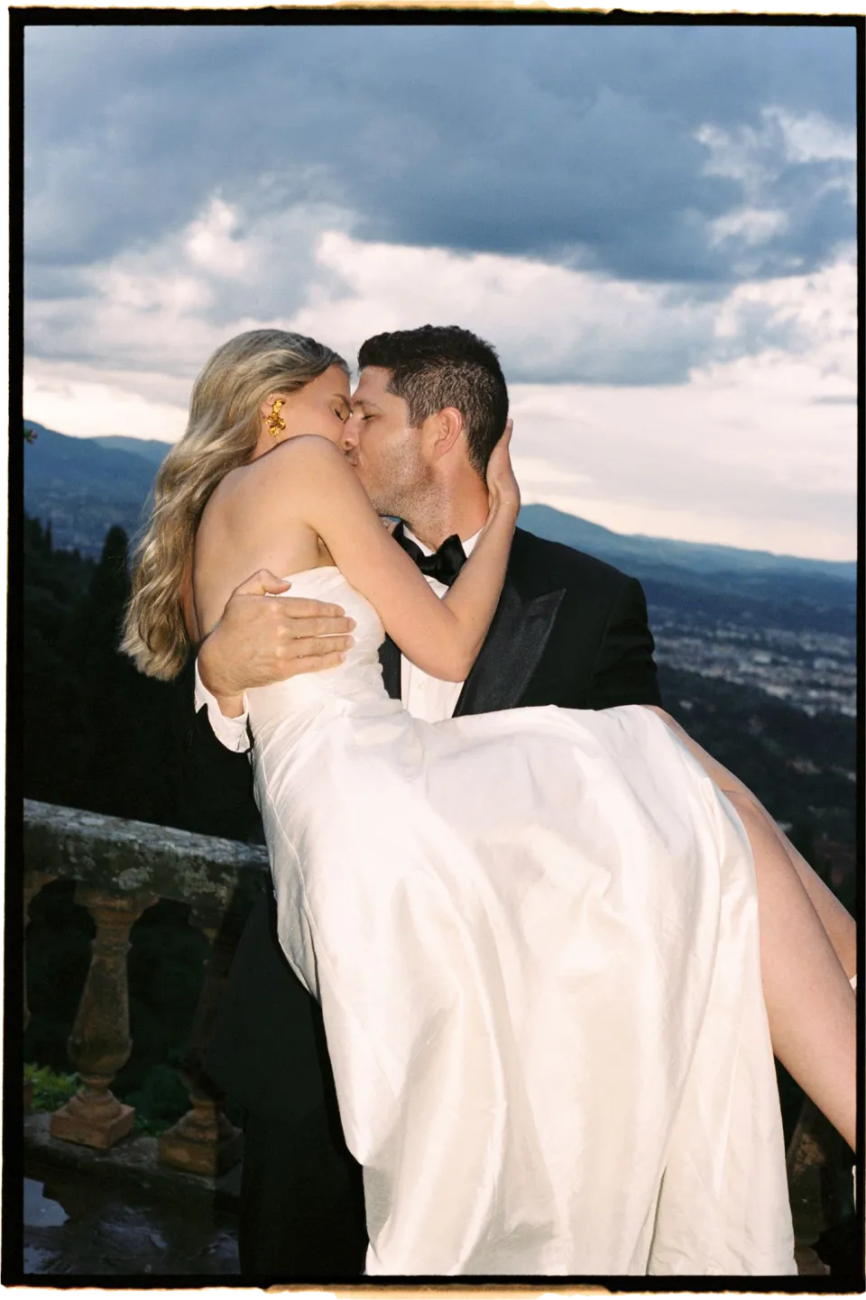 Holly and Matthew kissing as he holds her in his arms, lit by flash beneath the cloudy Tuscan sky, at Villa San Michele in Fiesole above Florence, Tuscany, Italy, for an Elopement Collective elopement. Film photograph by Khara from House of Lucie.