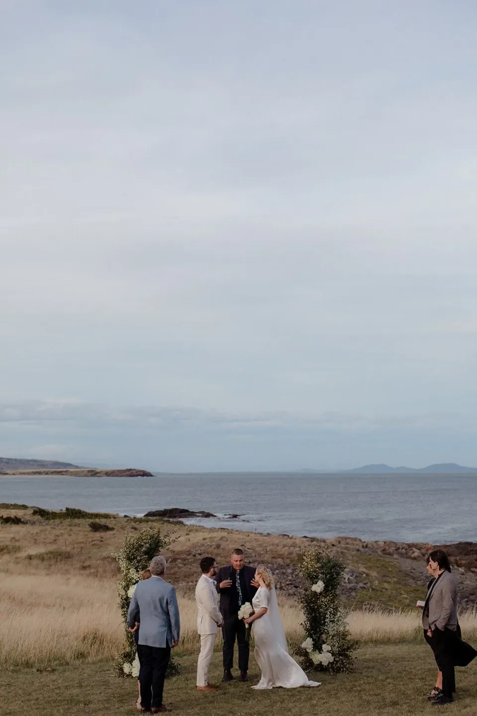 A small group gathers around a couple in coastal grass overlooking the sea.