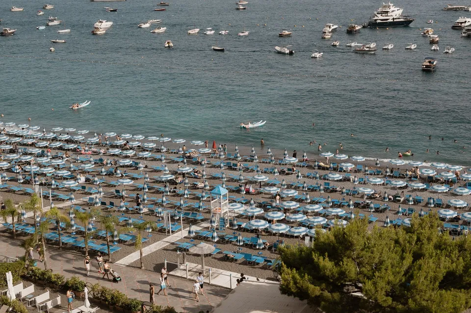 Rows of blue and white beach umbrellas and loungers at Spiaggia Grande, with swimmers and boats on the sea, in Positano on the Amalfi Coast, Italy, for an Elopement Collective elopement. Photograph by Jason Corroto.