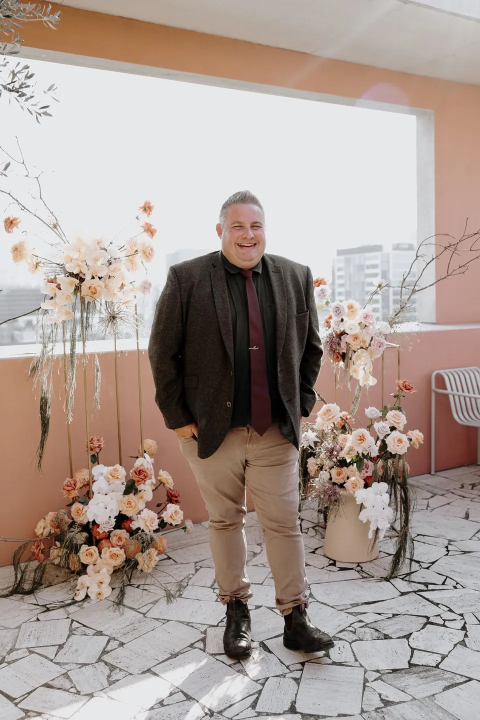 Celebrant Josh Withers standing between the terrace flower arrangements by Mrs Gibbons Florals with Brisbane behind him, at The Calile Hotel in Fortitude Valley, Brisbane, for an Elopement Collective elopement. Photo by George Bowden.