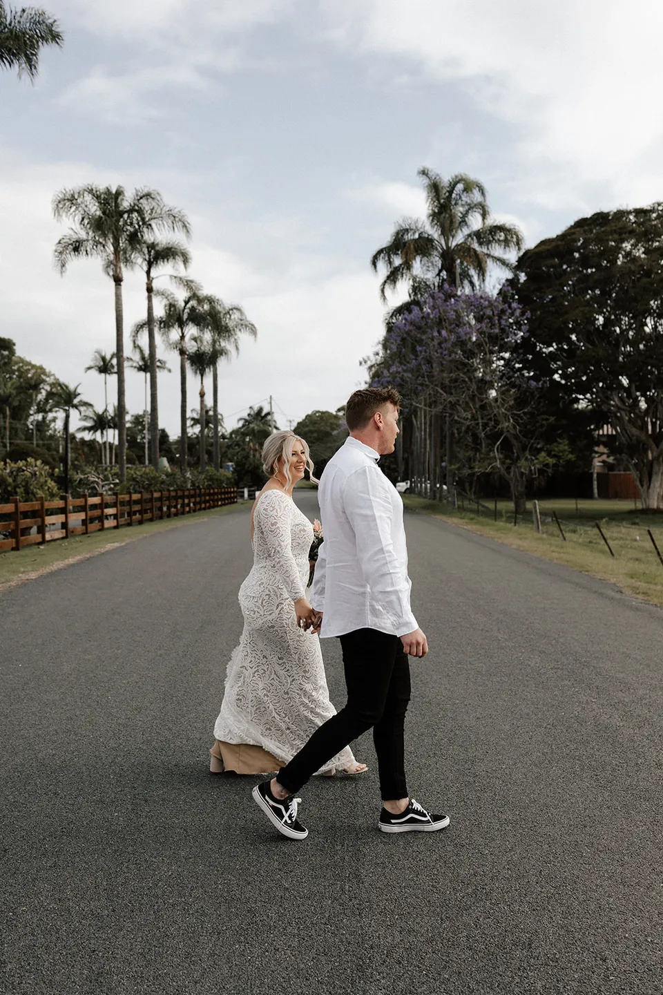 Matt and Laura walking hand in hand across a palm-lined country road, in the Tweed Valley, NSW, for an Elopement Collective elopement. Photo by Bulb Creative.