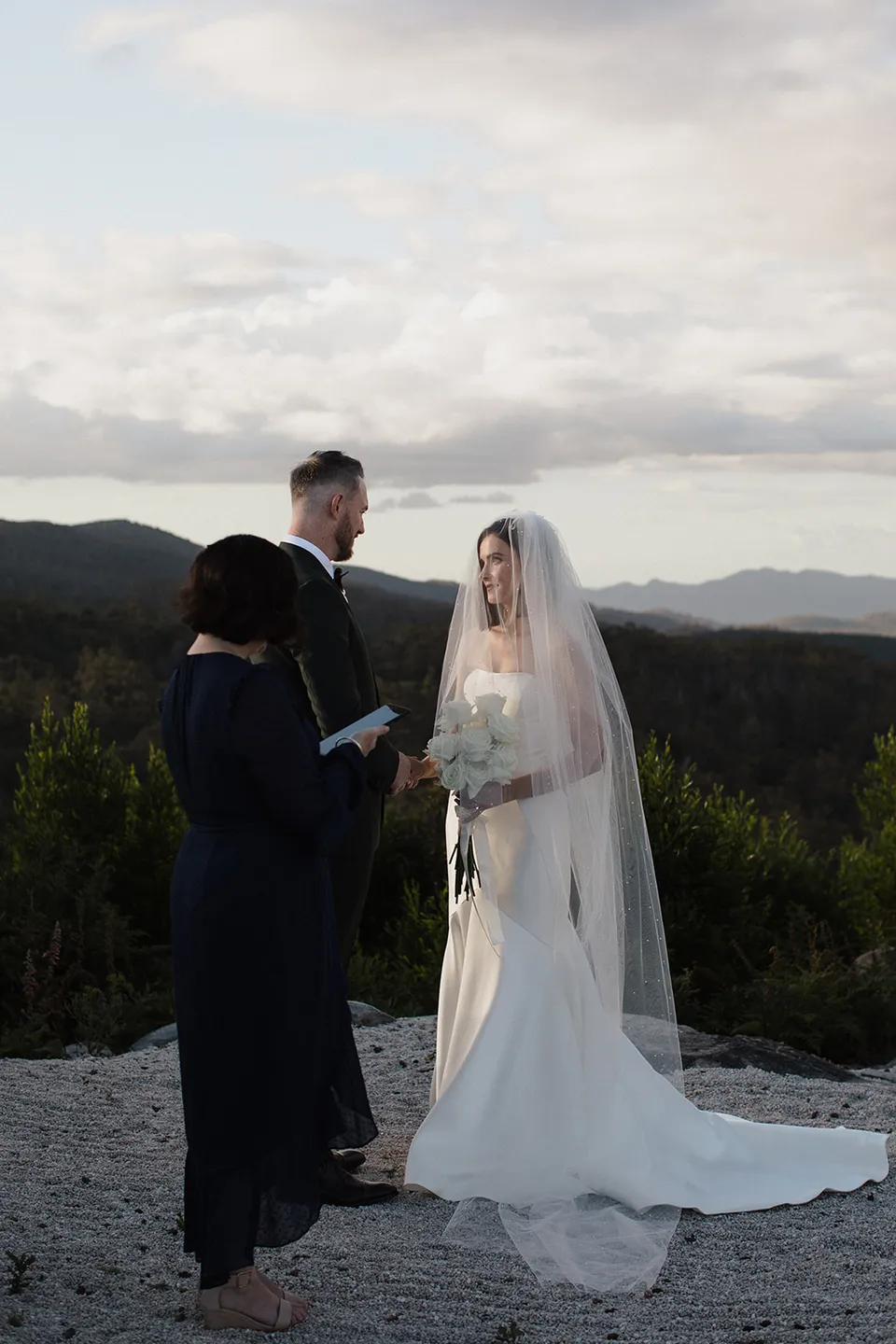 Celebrant Kath Tilly leading Meg and Ryan’s ceremony on the hilltop, with forest and distant ridges behind them. At The Keep on Tasmania’s east coast, for an Elopement Collective elopement. Photograph by Jenna Agius.