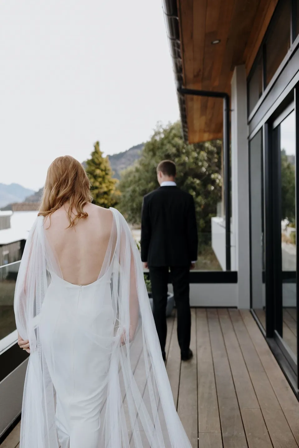 Neve approaching Hamish on a timber terrace for their first look, with mountains beyond the house, at their Airbnb in Fernhill, Queenstown, New Zealand, for an Elopement Collective Queenstown elopement. Photo by George Bowden.