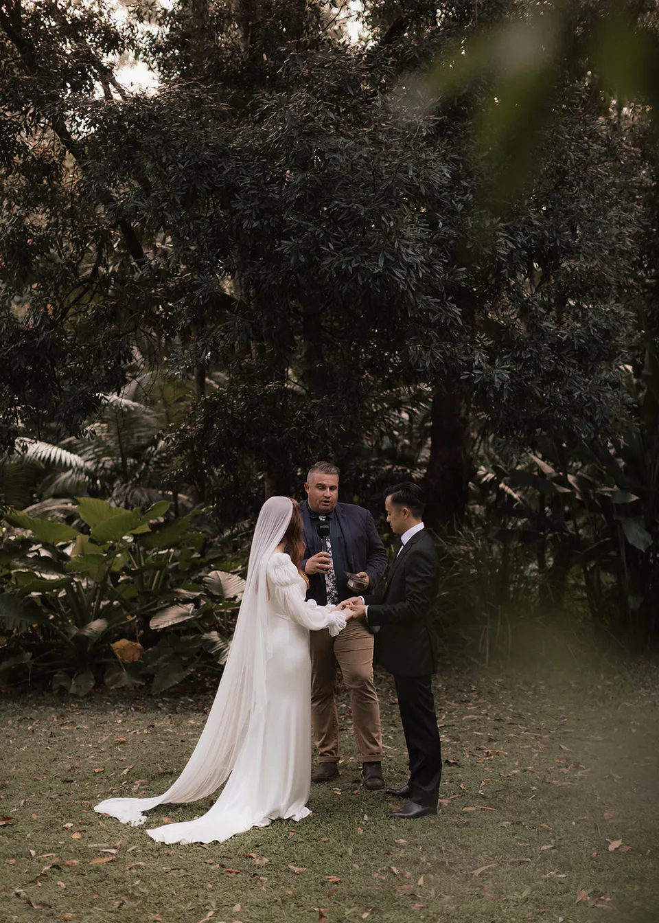 Elyse and Jonathan smiling with celebrant Josh Withers on the garden lawn. At Fig Tree Villa in Ewingsdale, Byron Bay, for an Elopement Collective elopement. Photograph by Bulb Creative.