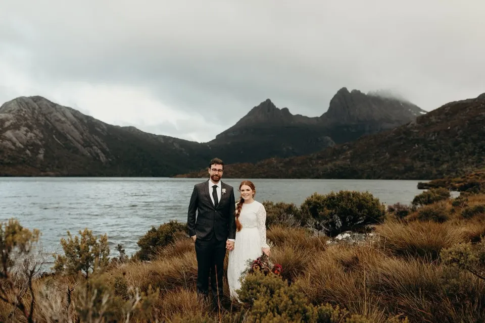 A couple stand beside a lake with rugged mountain peaks rising behind them.