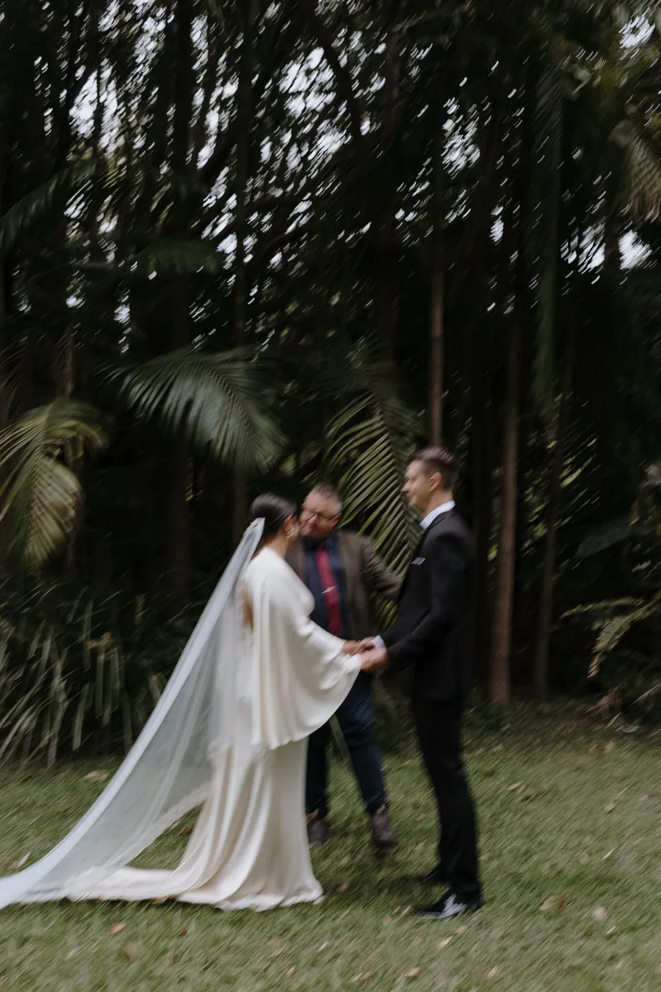 Laura and Matthew holding hands for their garden ceremony, her long veil trailing across the lawn. At Fig Tree Villa in Ewingsdale, Byron Bay, for an Elopement Collective elopement. Photograph by George Bowden.