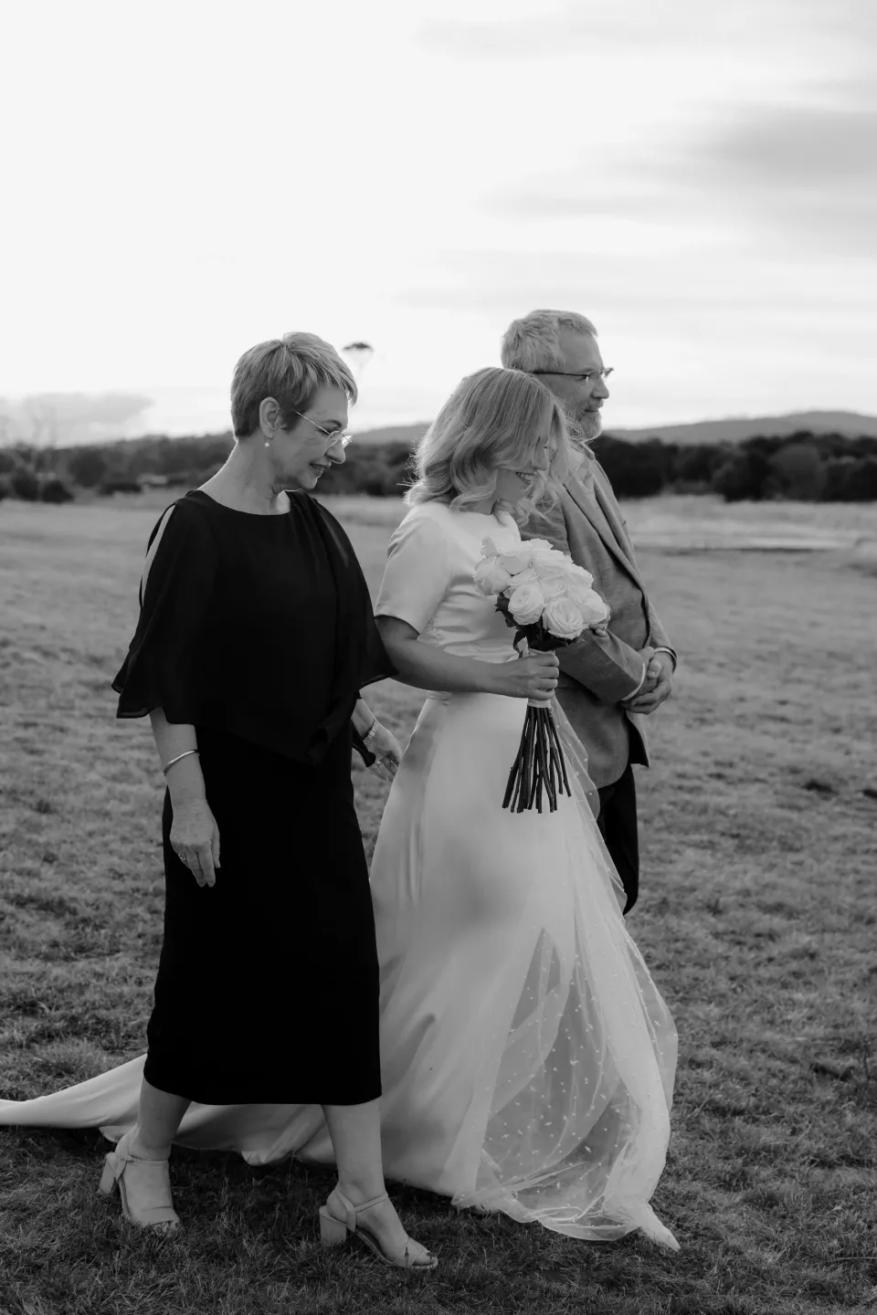 Black and white photograph of Jemmah and loved ones crossing the meadow, at Thalia Haven on the East Coast of Tasmania, Australia, for an Elopement Collective elopement. Photograph by Joey Willis.