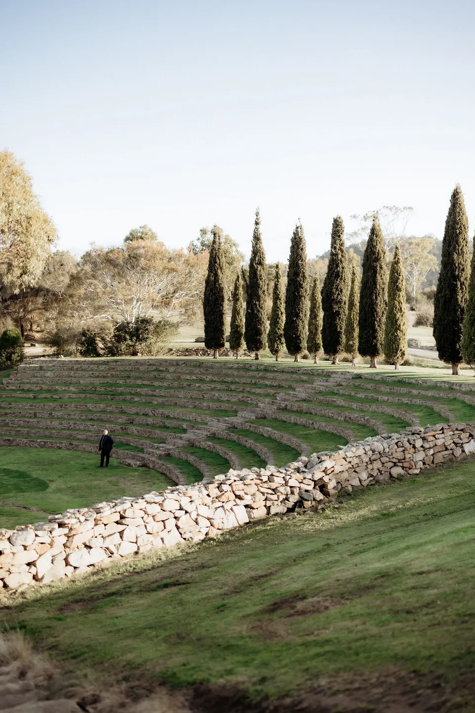 Tall cypress trees framing Piermont’s stone amphitheatre, with the celebrant waiting on the grass below. At Piermont Retreat on Tasmania’s east coast, for an Elopement Collective elopement. Photograph by Omar of By The Wilde.
