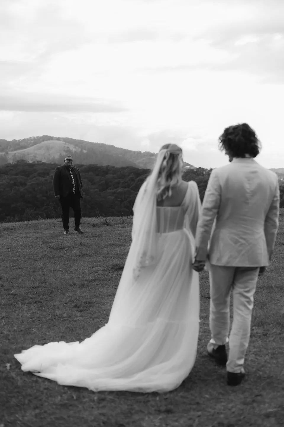 Black-and-white rear view of Bridget and James walking towards their outdoor ceremony. At Cudgera Creek in northern New South Wales, for an Elopement Collective elopement. Photograph by Bulb Creative.