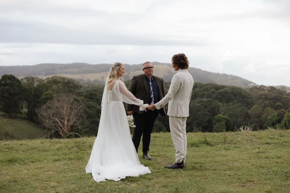 Bridget and James holding hands as Josh Withers leads their ceremony overlooking the hills. At Cudgera Creek in northern New South Wales, for an Elopement Collective elopement. Photograph by Bulb Creative.