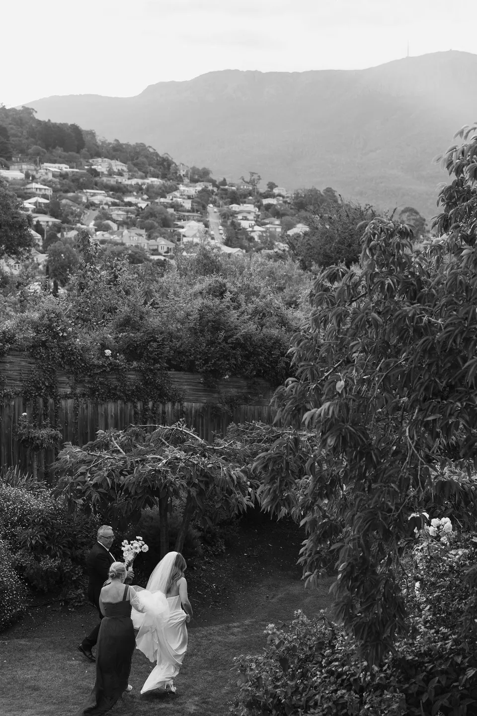 Black and white view across the hotel garden towards the hillside and mountain, with Tanesha approaching the ceremony below, at Islington Hotel in South Hobart, Tasmania, Australia, for an Elopement Collective summer elopement. Photograph by George Bowden.