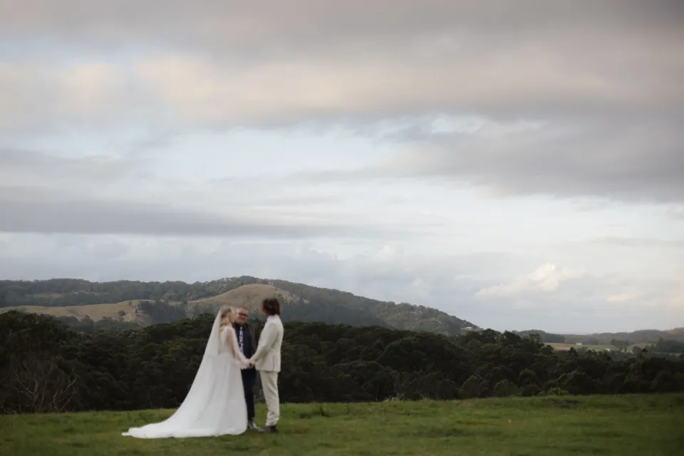 Wide view of Bridget and James's hillside ceremony beneath a cloudy sky. At Cudgera Creek in northern New South Wales, for an Elopement Collective elopement. Photograph by Bulb Creative.