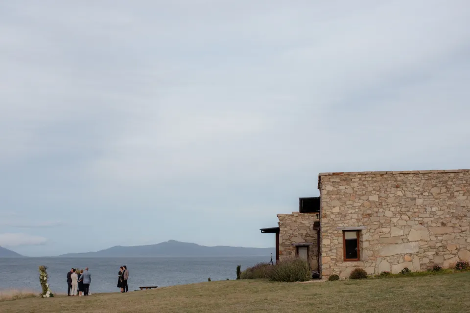 A wide view of the stone accommodation and wedding ceremony above the bay, at Thalia Haven on the East Coast of Tasmania, Australia, for an Elopement Collective elopement. Photograph by Joey Willis.
