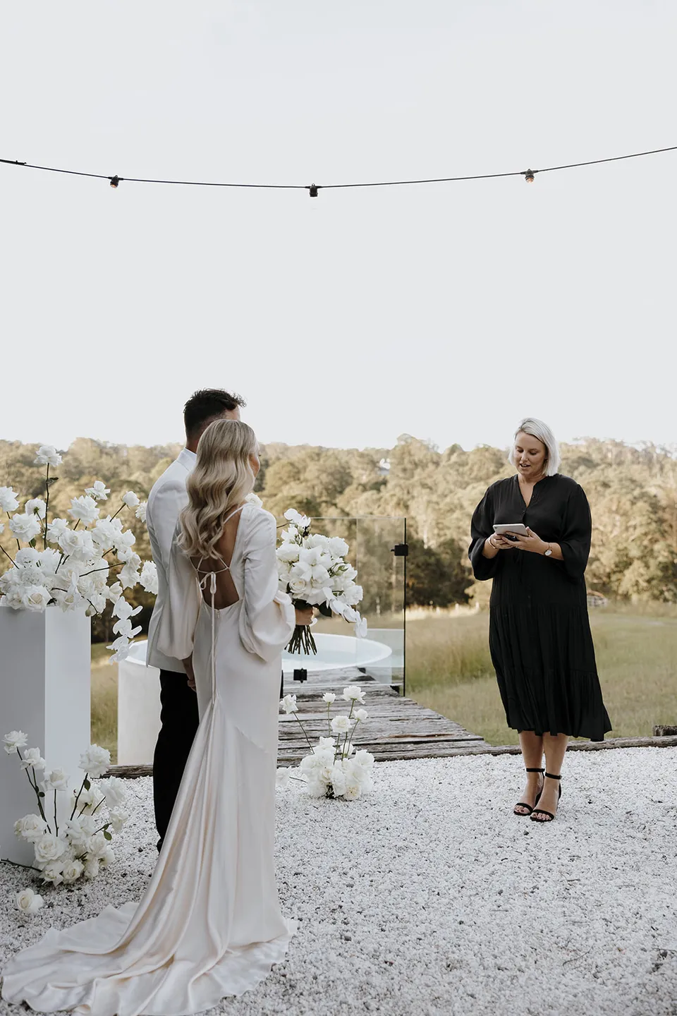 Hayley Hart leading Gabrielle and Greg’s outdoor ceremony between white floral plinths by Mrs Gibbons beneath festoon lights, at Roundhill Retreat in Obi Obi, Sunshine Coast Hinterland, Queensland, for an Elopement Collective elopement. Photo by George Bowden.