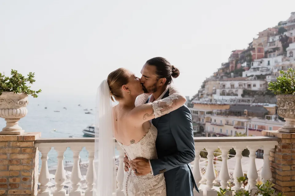 Skyler and Zach kissing beside the terrace balustrade, with the sea and Positano’s hillside houses behind them, at Hotel Buca di Bacco in Positano, Amalfi Coast, Italy, for an Elopement Collective elopement. Photograph by Jason Corroto.