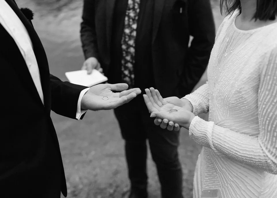 Black-and-white close-up of April and Josh’s open hands during the ceremony, with Josh Withers behind them, at Dove Lake, Cradle Mountain, Tasmania, for an Elopement Collective elopement. Photo by Bulb Creative.