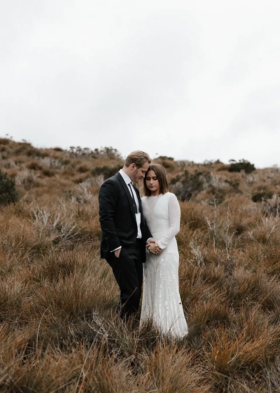 A couple embrace in tall, tawny grasses beneath an overcast sky.