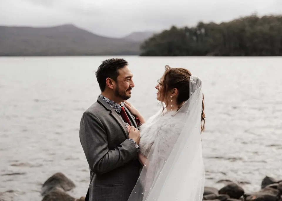 Sarah and Jono smiling at each other beside the lake, with mountains rising behind them, at Pumphouse Point, Lake St Clair, Tasmania, for an Elopement Collective elopement. Photo by Bulb Creative.