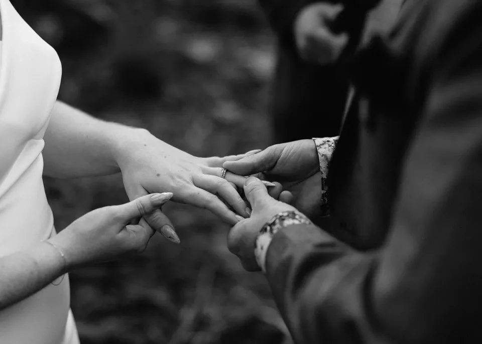 Black-and-white close-up of Jono placing a wedding ring on Sarah’s hand, at Pumphouse Point, Lake St Clair, Tasmania, for an Elopement Collective elopement. Photo by Bulb Creative.