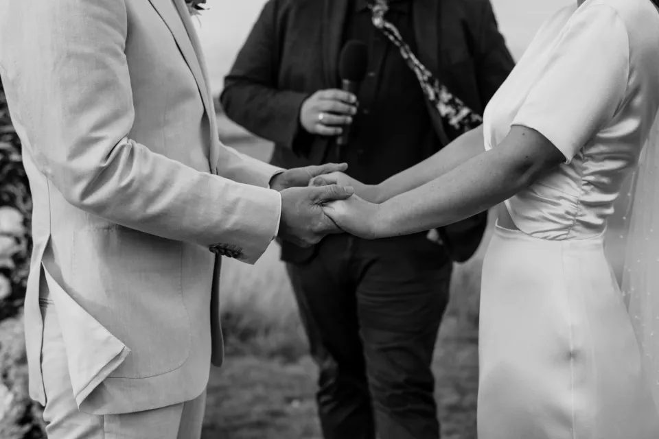 Black and white close-up of Jemmah and Jack holding hands beside Josh Withers, at Thalia Haven on the East Coast of Tasmania, Australia, for an Elopement Collective elopement. Photograph by Joey Willis.