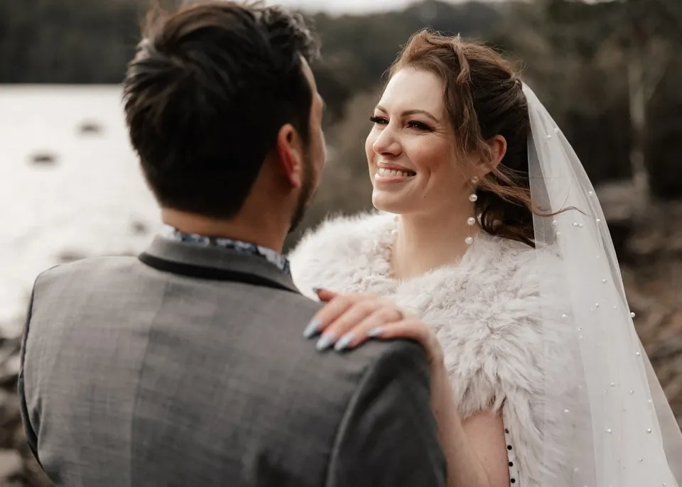 Sarah smiling at Jono with her hand resting on his shoulder and her veil gathered behind her, at Pumphouse Point, Lake St Clair, Tasmania, for an Elopement Collective elopement. Photo by Bulb Creative.