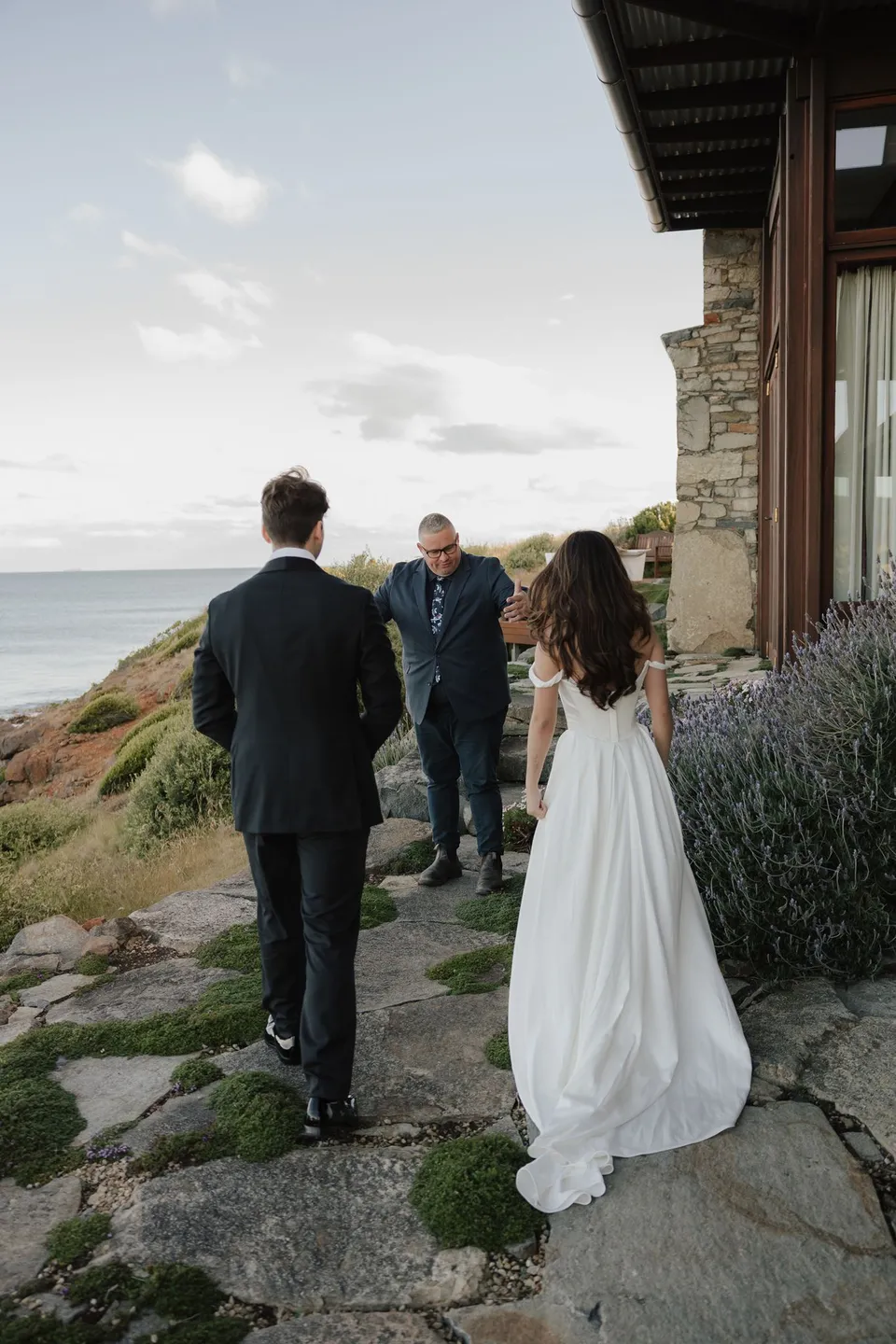 Rear view of Clara and Harry walking along a stone path towards celebrant Josh Withers beside the house, at Thalia Haven on the East Coast of Tasmania, for an Elopement Collective elopement. Photograph by Bulb Creative.