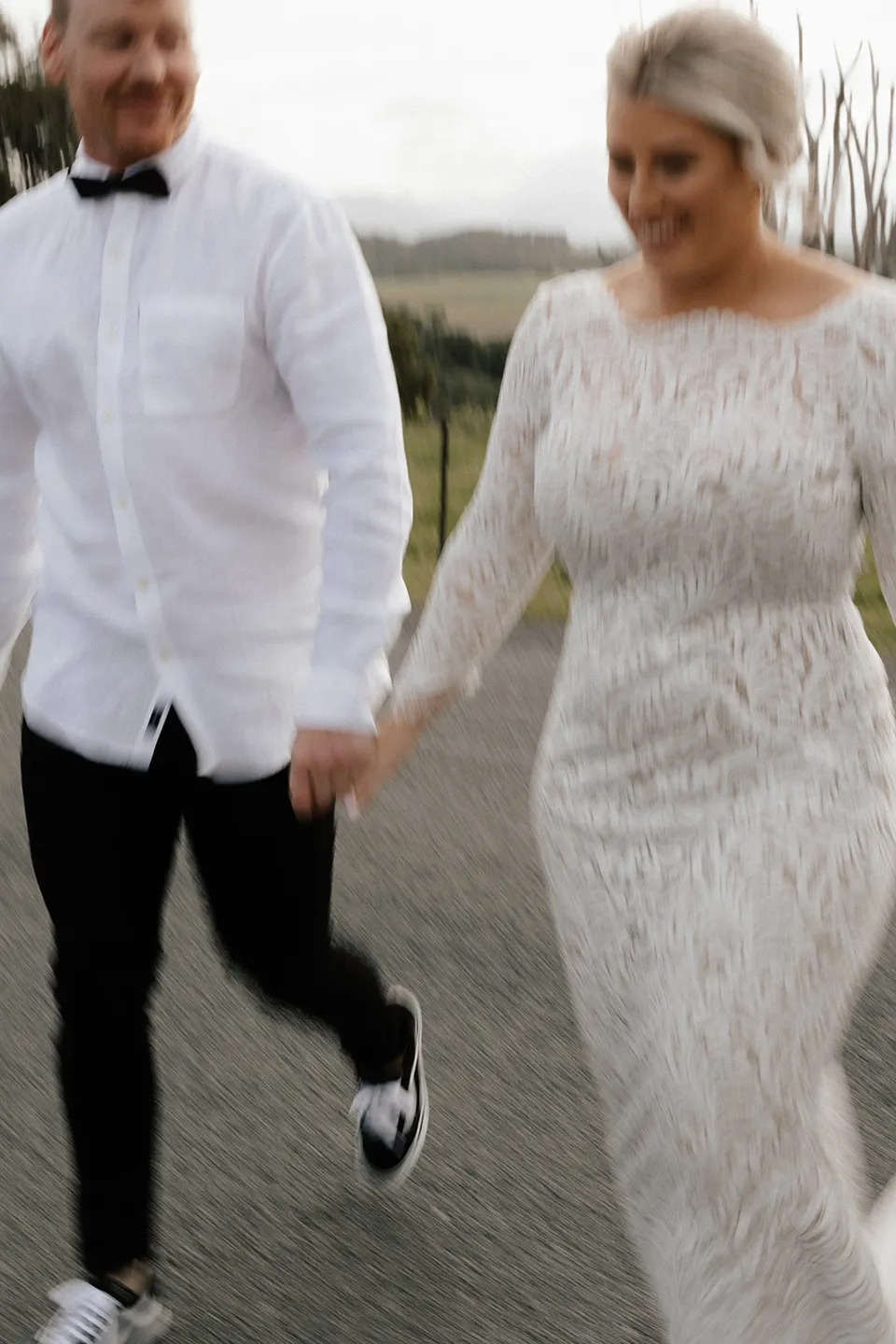 Matt and Laura holding hands as they walk, with Laura’s lace dress moving around her, in the Tweed Valley, NSW, for an Elopement Collective elopement. Photo by Bulb Creative.
