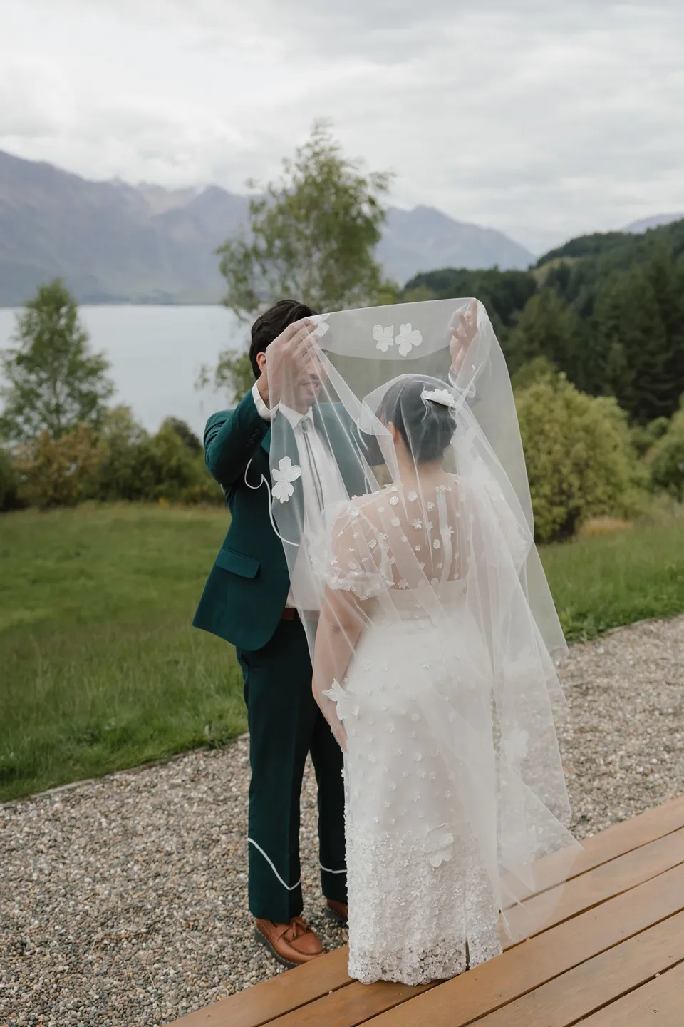 Adrian lifting Jaeda’s flower-appliqué veil during their first look, with the lake and mountains beyond the deck, at their alpine Airbnb in Ben Lomond, Queenstown, New Zealand, for an Elopement Collective Queenstown elopement. Photograph by Bulb Creative.